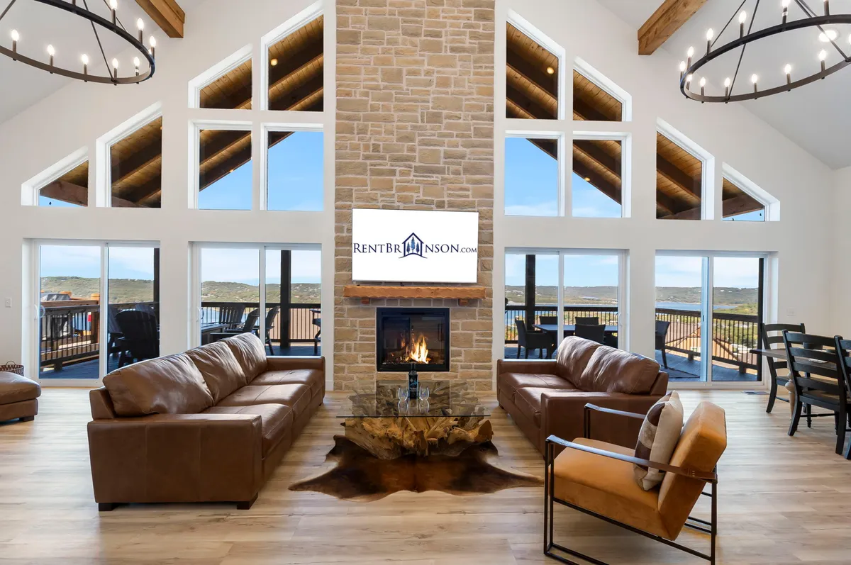 A spacious open-concept living area featuring a central stone fireplace, large comfortable leather sofas, rustic wooden beams, and a dining area positioned by expansive windows that offer sweeping views of Table Rock Lake.
