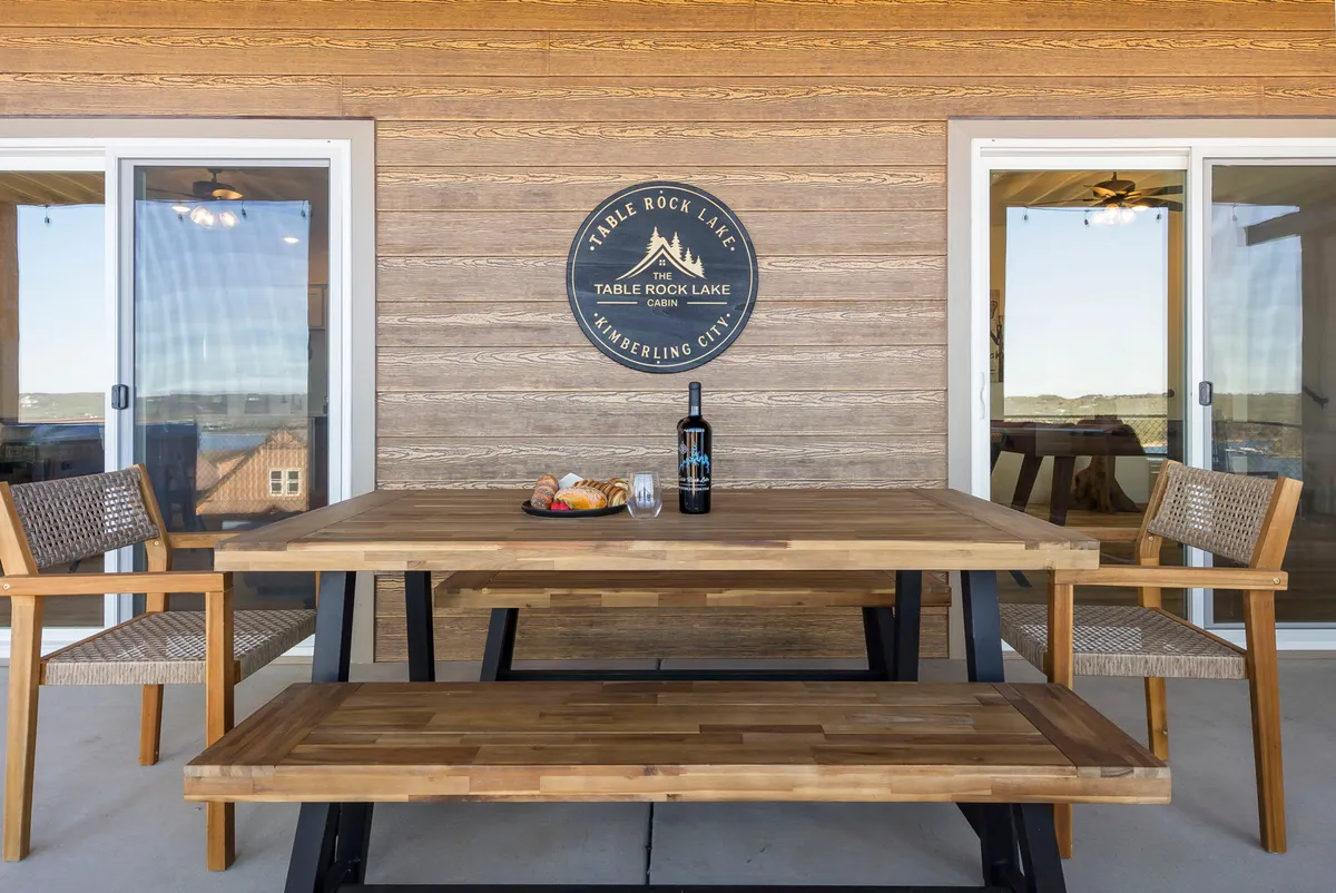 An outdoor dining setup with a wooden table adorned with a bottle of wine and baked goods, set against a stunning backdrop of Table Rock Lake. The scene is relaxed and welcoming, suggesting al fresco dining and celebration.