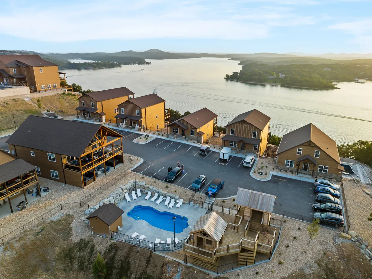 This image features a vibrant exterior scene showcasing a row of private cabins, a sparkling swimming pool, a playground structure, and a lake in the background. The space is bustling with potential for both relaxation and recreation, capturing the essence of a resort community.