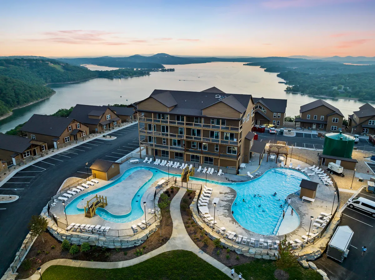 This image captures an inviting exterior scene featuring multiple swimming pools, landscaped gardens, a hotel building, and a stunning lake view in the background. The relaxing outdoor areas, lounge seating, and open sky evoke tranquility and a sense of retreat in nature.