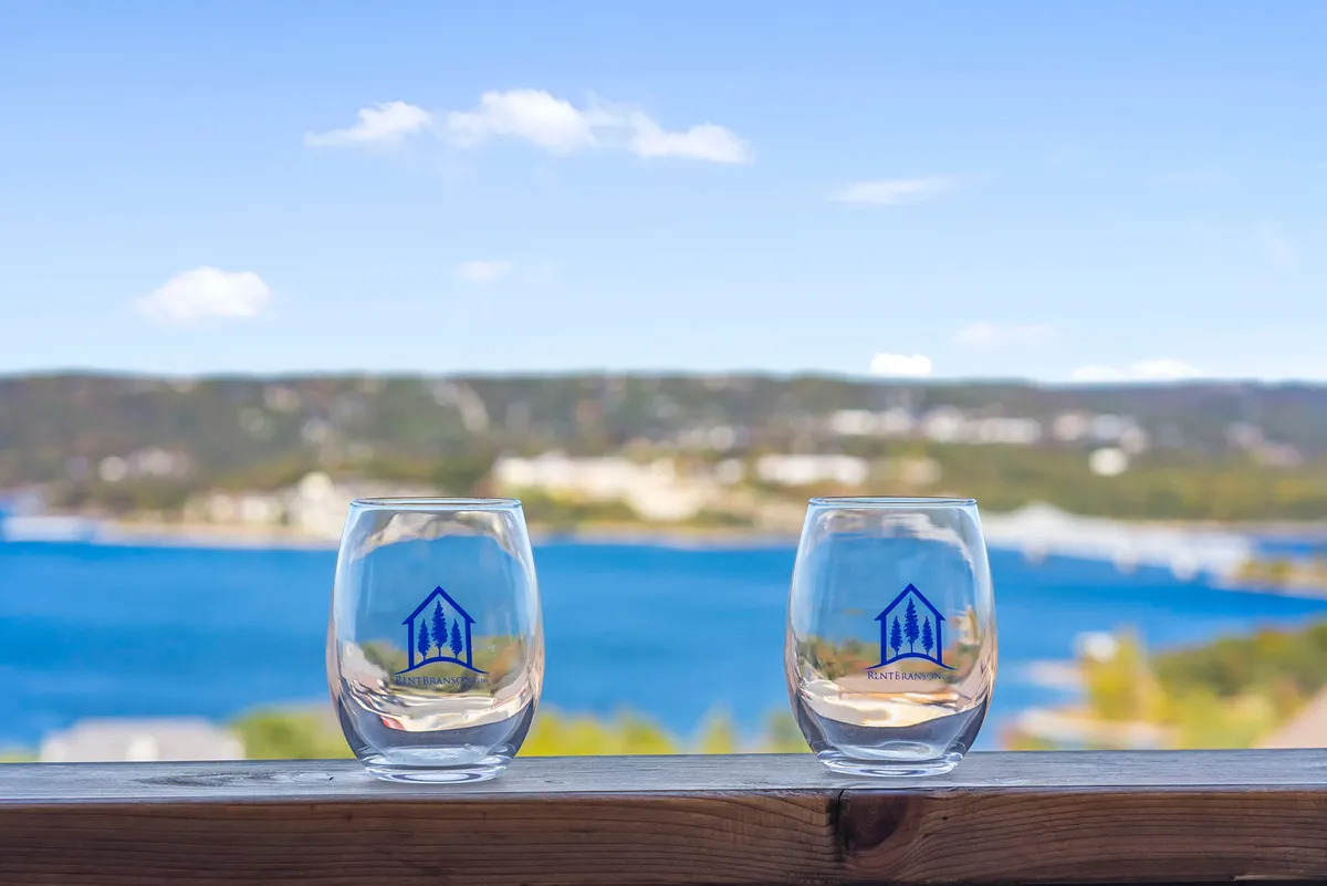 The image shows two branded glasses set on a terrace, with a sweeping view of rolling hills and glistening water in the distance. The scene is bathed in soft light, suggesting sunset or early evening, and evokes relaxation and togetherness while enjoying the landscape.