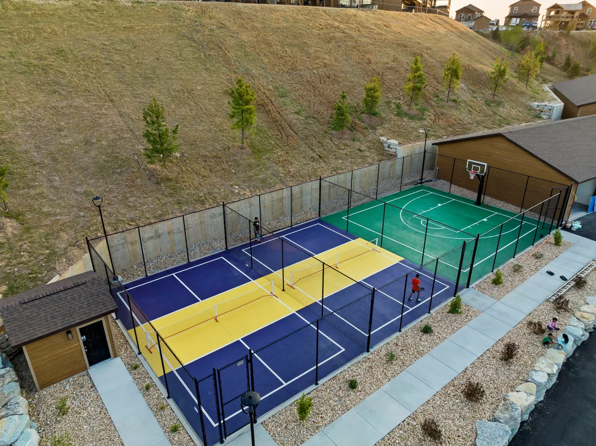 This vibrant exterior shot features both tennis courts and a basketball court surrounded by natural landscaping, with light fixtures and a storage shed in the background. The scene suggests active play and opportunities for friendly matches and community events.