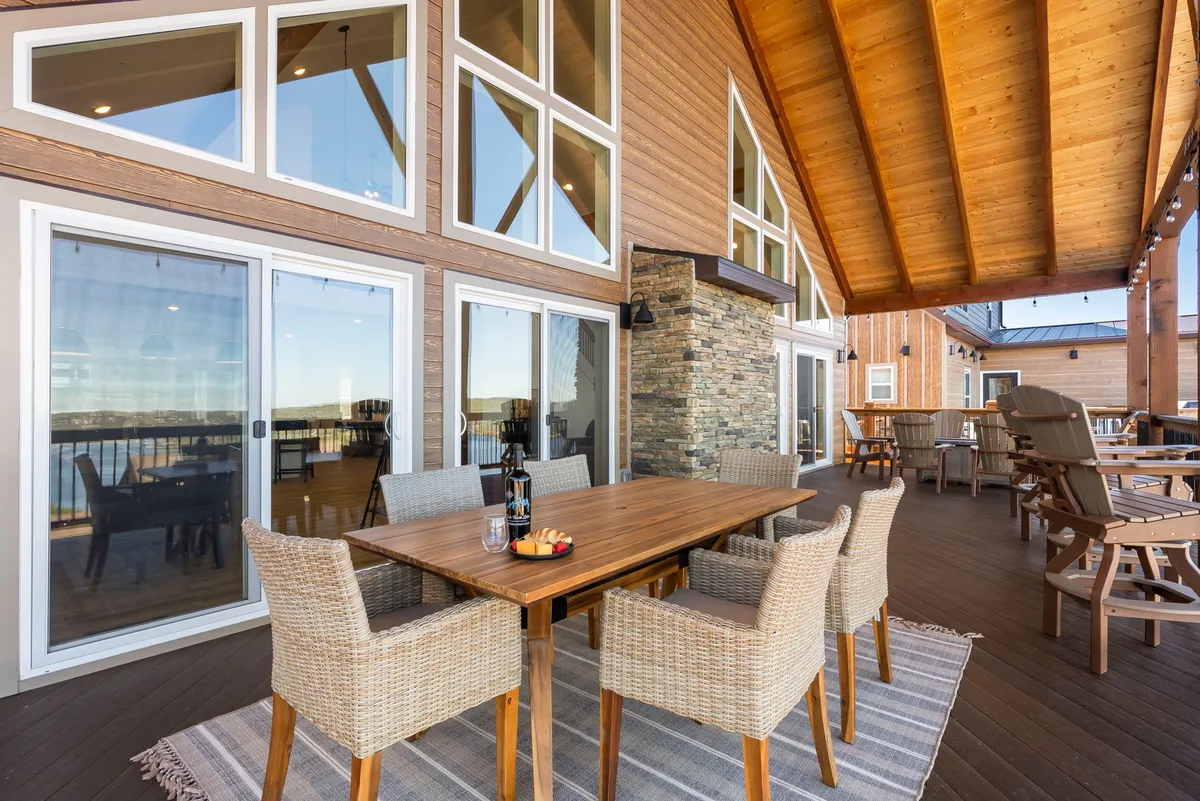 An exterior view featuring a large outdoor dining table set on a patio, with views through expansive windows and a decorative stone wall, suggesting a space for both gathering and relaxation. The inviting setup hints at outdoor meals, games, and casual socializing.