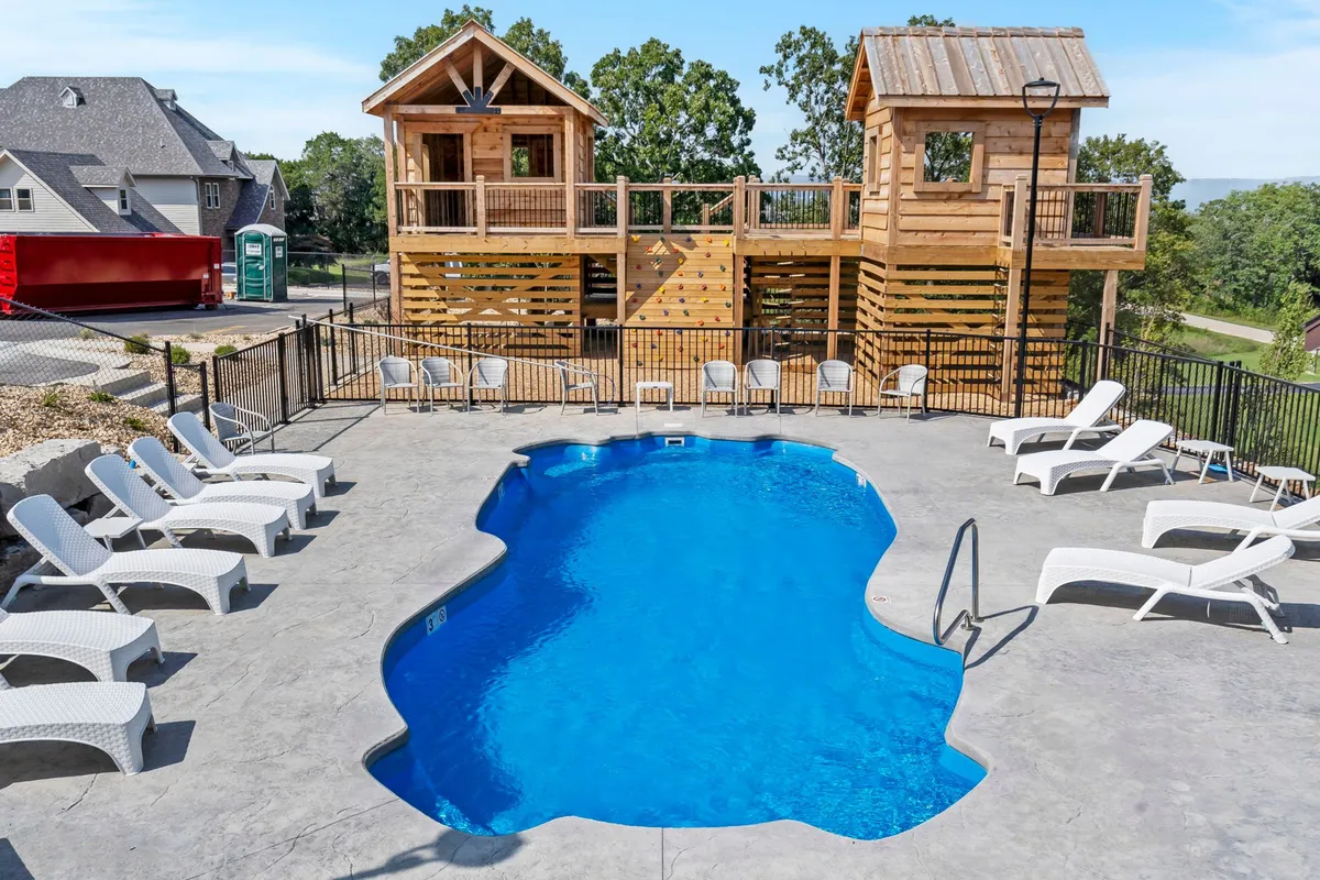 The photo presents an exterior view of a swimming pool surrounded by wooden playhouses and lounge chairs. Children can be imagined playing in the treehouse area while families unwind poolside, blending playful adventure with relaxation.