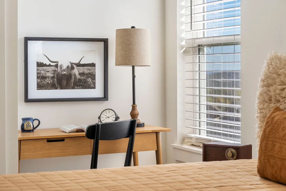 A cozy space featuring a desk with a clock and book, table lamp, window with blinds, and framed artwork of a cow above. The ambiance is conducive to quiet work or creative relaxation, making it ideal for the upper-level queen bedroom with a designated desk and inspiring views.