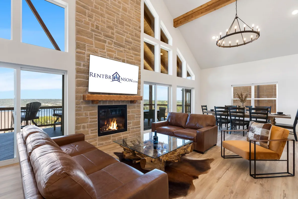 The image showcases a stunning great room with a soaring stone fireplace, exposed wooden beams, and expansive windows flooding the space with natural light and framing beautiful lake views. A sleek glass coffee table anchors a plush seating area, inviting guests to relax and socialize while soaking up the warmth and scenery.