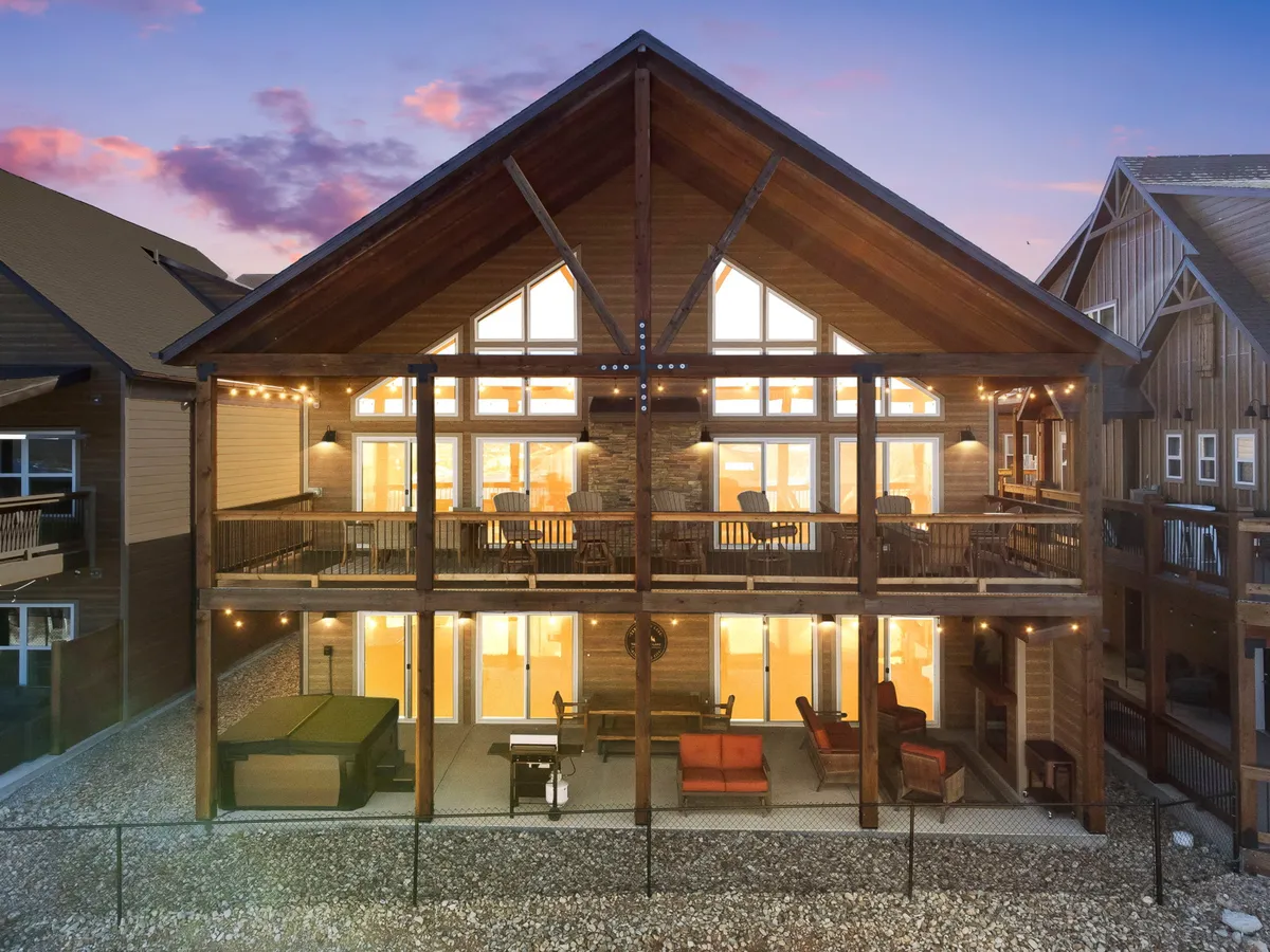 This image features a large wooden deck attached to a private home, complete with outdoor seating areas, a hot tub, and warmly lit windows. The inviting deck overlooks scenic surroundings, offering a serene spot to unwind.