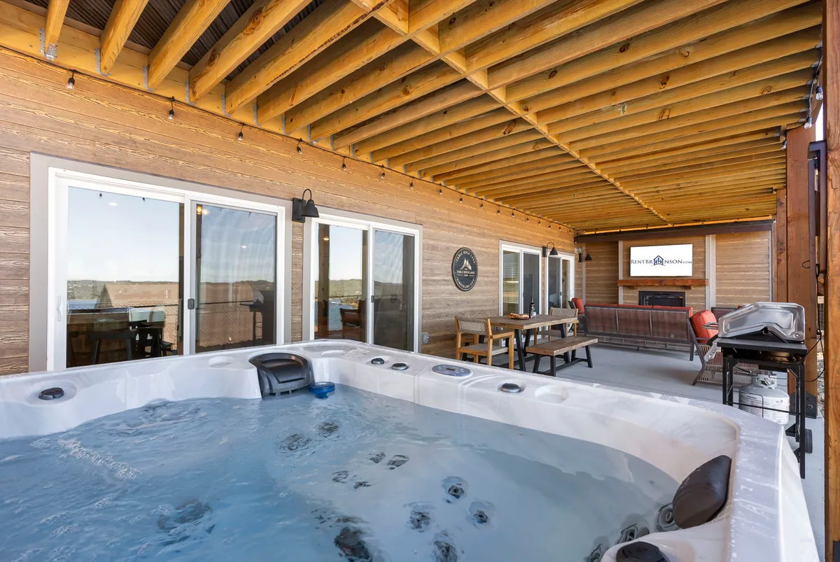 A spacious, covered outdoor patio with a private hot tub, outdoor seating, dining table, and a modern fireplace. Large sliding glass doors connect the indoor entertainment space to the outdoor retreat, blending indoor and outdoor fun.