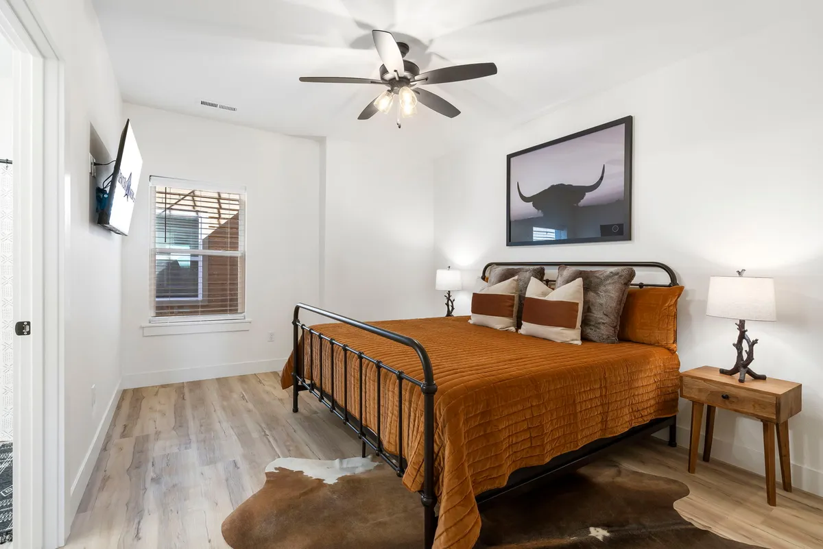 A king bedroom with orange bedding, artwork featuring a bull's head above the bed, wooden bedside tables, and a cowhide rug on the floor. The space is flooded with natural light, creating a warm and welcoming atmosphere that reflects western decor and a focus on comfort.